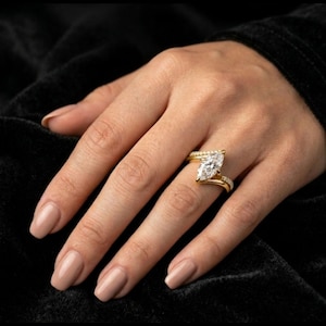 May include: A gold engagement ring with a marquise-cut diamond is displayed on a hand. The ring has a delicate band with small accent stones. The hand has a neutral nail polish color and is set against a black background.
