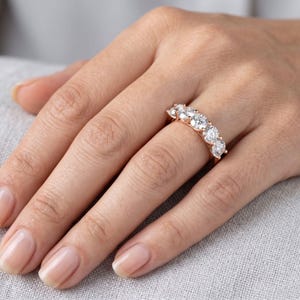 Könnte beinhalten: Ein Ring aus Roségold mit fünf klaren, rund geschliffenen Steinen in einer Reihe. Der Ring wird an einem Finger getragen und zeigt das metallische Band sowie die facettierten, funkelnden Steine, die horizontal angeordnet sind.