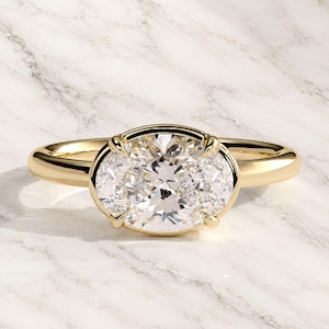 May include: A gold ring featuring a large, horizontally oriented oval-cut clear gemstone set in a four-prong setting. The band is smooth and polished, crafted from a yellow-toned metal that complements the brilliant, faceted center stone.