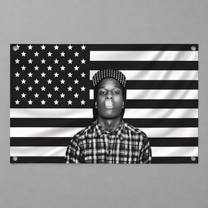 Op de afbeelding: Een rechthoekige wandbanner met een zwart-wit afbeelding van een persoon met een pet met patroon en een geruit overhemd. De achtergrond toont een zwart-wit Amerikaans vlagontwerp met metalen ringen in de hoeken voor bevestiging.