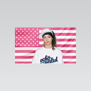 Peut inclure: Un t-shirt blanc avec le texte bleu ecko unltd imprimé sur le devant, porté avec une casquette de baseball blanche à bord sombre. L'arrière-plan est un drapeau avec des rayures horizontales roses et blanches et des étoiles blanches sur fond rose.