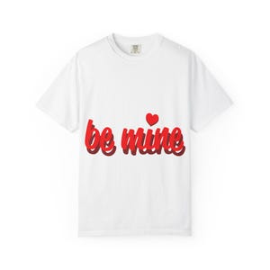Op de afbeelding: Wit T-shirt met de tekst "be mine" in rode letters, vergezeld van een klein rood hart. De tekst heeft een schaduweffect, wat diepte aan het ontwerp toevoegt. Een eenvoudig, romantisch grafisch T-shirt.
