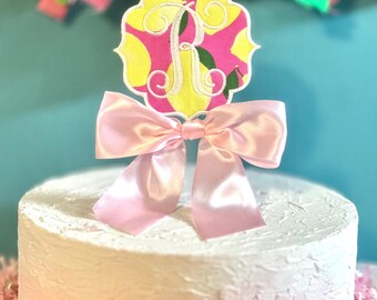 May include: A cake topper featuring a quatrefoil-shaped plaque with a yellow lemon and green leaf pattern on a pink background, centred with a white embroidered letter R. A light pink satin ribbon bow is attached to the base of the plaque.