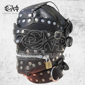 Op de afbeelding: Een zwart lederen volledig hoofdmasker met zilverkleurige metalen studs, verstelbare gespriemen en een horizontale ritssluiting bij de mond, beveiligd met een klein messing hangslot. Een grote metalen ring is bevestigd aan de halsband.