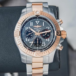 May include: A Breitling chronograph watch with a silver and rose gold case and band. The watch face is gray with black sub-dials and rose gold hands and markers. The bezel has a rose gold finish with black numerals. The watch is displayed on a dark surface.