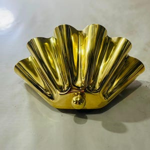 May include: A decorative, gold-colored metal bowl in the shape of a seashell. The bowl has a scalloped edge and a rounded base with a small, spherical detail. The surface is highly reflective.