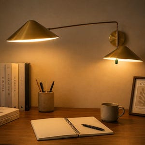 Könnte beinhalten: Eine Wandleuchte aus Messing mit zwei konischen Schirmen an einem schwenkbaren Arm. Die Lampe strahlt ein warmes, nach unten gerichtetes Licht aus und beleuchtet einen Schreibtisch mit Notizbuch, Stift, Tasse, Stiftehalter, Büchern und Bild.