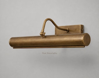 May include: A wall-mounted picture light fixture crafted from antique brass with a brushed finish. The design features a rectangular backplate, a curved adjustable arm, and a long cylindrical light housing. The text Noor BrassLights is visible.