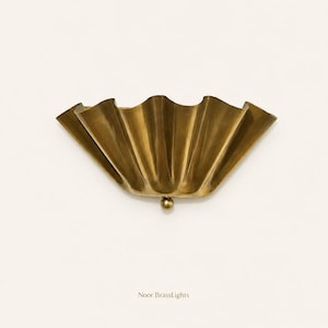 May include: A brass wall sconce with a scalloped, fan-like design. The sconce has a golden finish and a small, round detail at the bottom. The text "Noor BrassLights" is printed below the sconce.