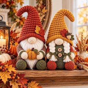 May include: Two crochet gnomes featuring autumn colors. The left gnome wears a rust-colored hat with a maple leaf and holds a small pumpkin. The right gnome wears a mustard-yellow hat with an acorn and leaf, featuring white braided hair.