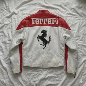 May include: White leather jacket with red accents and the Ferrari logo across the back shoulders. A black prancing horse emblem is centered below the logo. The jacket has a classic motorcycle style.