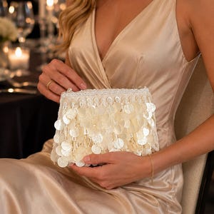 Peut inclure: Une pochette rectangulaire recouverte de sequins circulaires blancs irisés superposés et de petites perles blanches. Le bord supérieur présente une rangée de perles blanches, et le sac possède une structure rigide avec un fermoir.