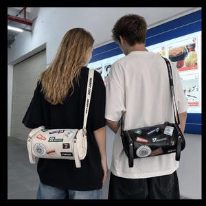 May include: Two cylindrical crossbody bags, one white and one black, each featuring various colorful rectangular and circular patches with text including "ST RETICHEN" and "FASHION," and a long adjustable shoulder strap with printed text.