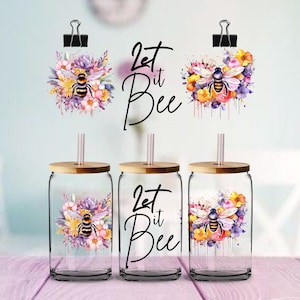 May include: Three glass tumblers with bamboo lids and straws, each featuring a watercolor design of a bee surrounded by flowers. The text "Let it Bee" is displayed on the center tumbler. The design includes purple, yellow, and pink flowers.