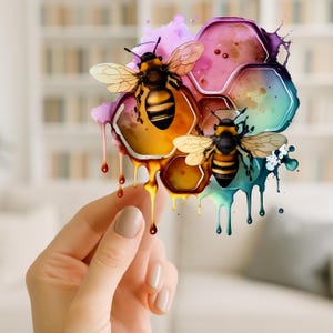 May include: A colorful watercolor illustration of two bees on a honeycomb. The honeycomb is a mix of pink, yellow, and blue hues, with dripping paint effects. The bees are black and yellow, with detailed wings. The image is held by a hand.