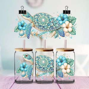 May include: Three clear glass tumblers with bamboo lids and straws, each featuring a printed design of a sea turtle with a teal and blue mandala shell, surrounded by blue and white hibiscus flowers, green leaves, seashells, and small pearls.