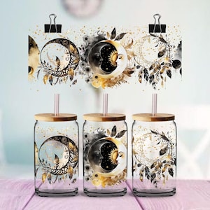 Puede incluir: Tres vasos de vidrio con tapas de madera y pajitas, con diseños celestiales de lunas, soles y plumas en oro, negro y blanco. Los vasos se exhiben con obras de arte a juego sujetas con clips.