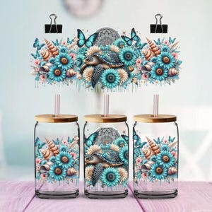May include: Three clear glass tumblers with bamboo lids and straws. Each tumbler has a watercolour design of a sea turtle, blue flowers, seashells, and butterflies. The design is repeated on a decorative element above the tumblers.