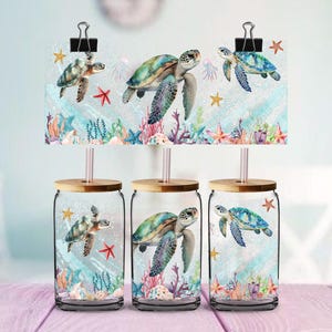 May include: Three glass tumblers with bamboo lids and straws, each featuring a watercolour design of sea turtles, starfish, and coral. The design is also printed on a card held by binder clips above the glasses. The tumblers are on a pink surface.