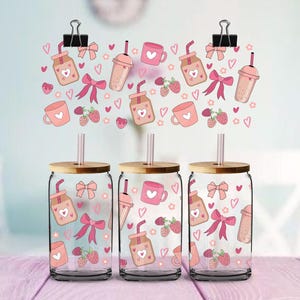 Könnte beinhalten: Drei transparente Trinkgläser mit Bambusdeckeln und Strohhalmen. Jedes Glas hat ein rosa-rotes Design aus Herzen, Schleifen und Getränken. Der Hintergrund zeigt ein ähnliches Muster, das von Büroklammern gehalten wird.