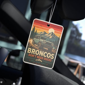 Peut inclure: Désodorisant rectangulaire avec un design vintage représentant un SUV bleu clair traversant un paysage de montagne sous un coucher de soleil. Le texte indique "BRONCOS DON'T NEED DUCKS" et "TRAIL BADGE SUPPLY EST. 2023".