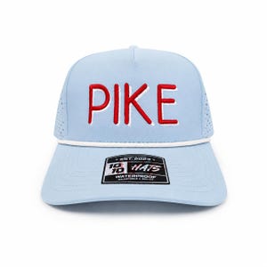 以下が含まれることがあります： 赤色の「PIKE」の文字が白い縁取りで刺繍された、水色の帽子。白いロープのディテールと「18 Hats」と書かれた白黒のタグが付いています。この帽子は防水で、調節可能です。