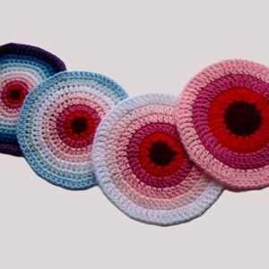 May include: Four colorful, crocheted coasters. Each coaster features concentric circles of color, with a dark center and a gradient of colors radiating outwards. Colors include blue, pink, red, and white. The coasters are round and appear handmade.