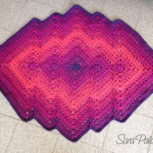May include: A crocheted diamond-shaped rug with a gradient of pink, purple, and dark purple colors. The rug has a scalloped edge and is made with a textured stitch.