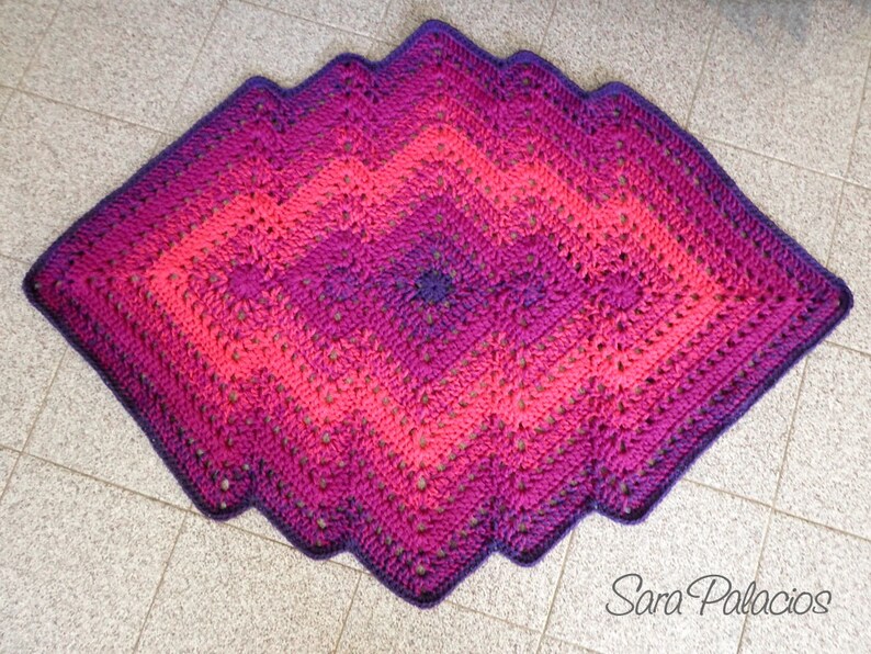 May include: A crocheted diamond-shaped rug with a gradient of pink, purple, and dark purple colors. The rug has a scalloped edge and is made with a textured stitch.