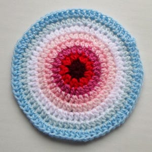 Può includere: Un sottobicchiere circolare fatto a mano, all'uncinetto con una sfumatura di colori. Il centro è rosso scuro, che sfuma attraverso tonalità di rosa, bianco e azzurro fino a un bordo esterno azzurro cielo. Ideale per l'arredamento della casa.