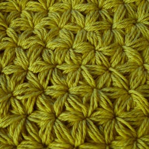 May include: Close-up of a handmade crochet fabric in a mustard yellow color. The texture features a repeating pattern of raised, star-shaped stitches, creating a dense and textured surface. This textile is suitable for various craft projects.