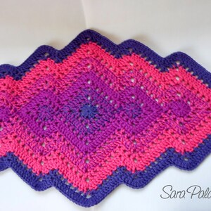 May include: A crocheted diamond-shaped rug with a purple and pink ombre design. The rug has a scalloped edge.
