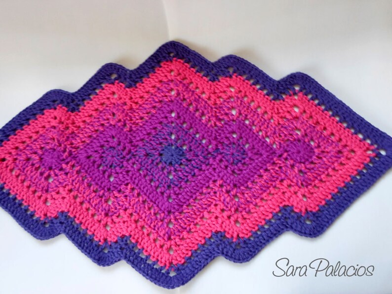 May include: A crocheted diamond-shaped rug with a purple and pink ombre design. The rug has a scalloped edge.