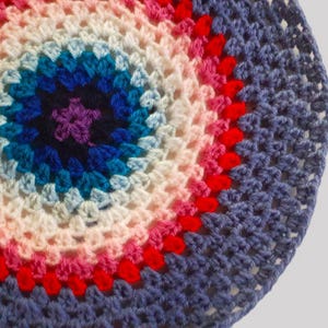 May include: A crocheted circular item with a vibrant, concentric design. The center features a purple star, surrounded by rings of blue, teal, white, pink, and red, set against a blue background. The handmade piece is likely a coaster or decorative item.