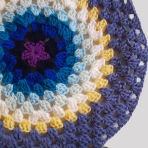Puede incluir: Un artículo circular tejido a ganchillo con un diseño floral. El centro presenta una flor morada, rodeada de anillos concéntricos azules, blancos y amarillos. El borde exterior es azul oscuro. La pieza hecha a mano es probablemente un posavasos o un elemento decorativo.