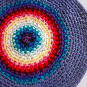 Puede incluir: Un artículo circular, tejido a ganchillo, con un vibrante diseño de arcoíris en el centro. El diseño presenta círculos concéntricos de color, incluyendo rojo, amarillo, azul y morado, sobre un fondo azul bígaro. El artículo es hecho a mano.