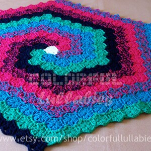 Puede incluir: Una manta de bebé de ganchillo colorida con un patrón en espiral en tonos de rosa, azul, verde y azul marino. La manta está hecha con un punto texturizado y tiene un centro blanco.