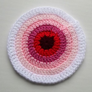 Puede incluir: Un posavasos circular de ganchillo hecho a mano con un diseño concéntrico. El posavasos presenta un anillo exterior blanco, seguido de anillos de rosa claro, rosa medio, rosa oscuro, rojo y un centro granate oscuro.