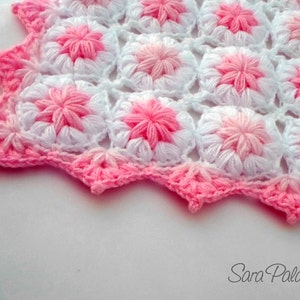 May include: A pink and white crocheted blanket with a scalloped edge. The blanket is made with a repeating flower pattern.