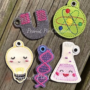 May include: Five felt science-themed keychains. The keychains are shaped like a lightbulb, a beaker, a DNA strand, a horseshoe magnet, and an atom. The lightbulb and beaker have smiling faces. The keychains are all different colors.