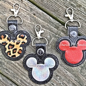 May include: Three black keychains with a Mickey Mouse head design. The keychains have different fabric inserts: leopard print, red, and iridescent.