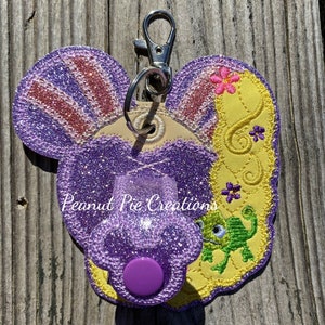 May include: Purple and yellow glitter keychain with a cartoon chameleon and a purple button. The keychain has the text "Peanut Pie Creations" on it.