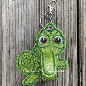 May include: A green glittery frog-shaped keychain with brown eyes and a silver clasp. The keychain has a loop and a strap with a snap closure. The text "Peanut Pie Creations" is printed on the strap.