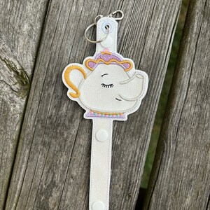 May include: White teapot with a purple lid and a sleepy face, attached to a white fabric strap with a silver key ring. The strap has three white buttons.