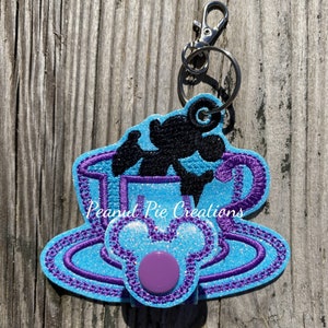 May include: A blue and purple glitter teacup keychain with a black silhouette of Mickey Mouse inside the cup. The keychain has a purple button and a silver metal ring and clip.