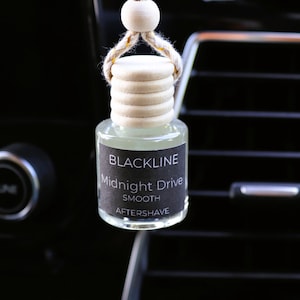 May include: A small, clear glass bottle of "Midnight Drive Smooth Aftershave" car air freshener. The bottle has a wooden cap and a string for hanging. The label is black with white text. The background is a car's dashboard and air vent.