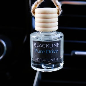 Może przedstawiać: Przezroczysty szklany odświeżacz powietrza do samochodu z drewnianą górą i sznurkiem. Etykieta głosi "BLACKLINE Pure Drive FRESH LINEN". Odświeżacz powietrza wisi przed kratką wentylacyjną samochodu.