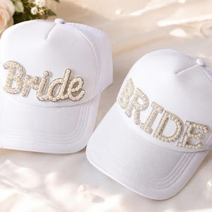 May include: Two white trucker-style caps featuring the word Bride in large, three-dimensional lettering. The letters are embellished with small, round, cream-colored faux pearls and clear crystals, creating a textured, sparkling appearance on the front.