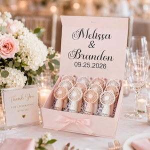 May include: A light pink rectangular gift box containing multiple small handheld fans, each personalized with the names Melissa and Brandon. The box lid features the text Melissa & Brandon 09.25.2026 in black script and sans-serif font.