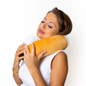 May include: A yellow and white neck pillow. The cylindrical pillow is held by a person. The pillow appears to be made of a soft, plush material. The person is wearing a white vest.